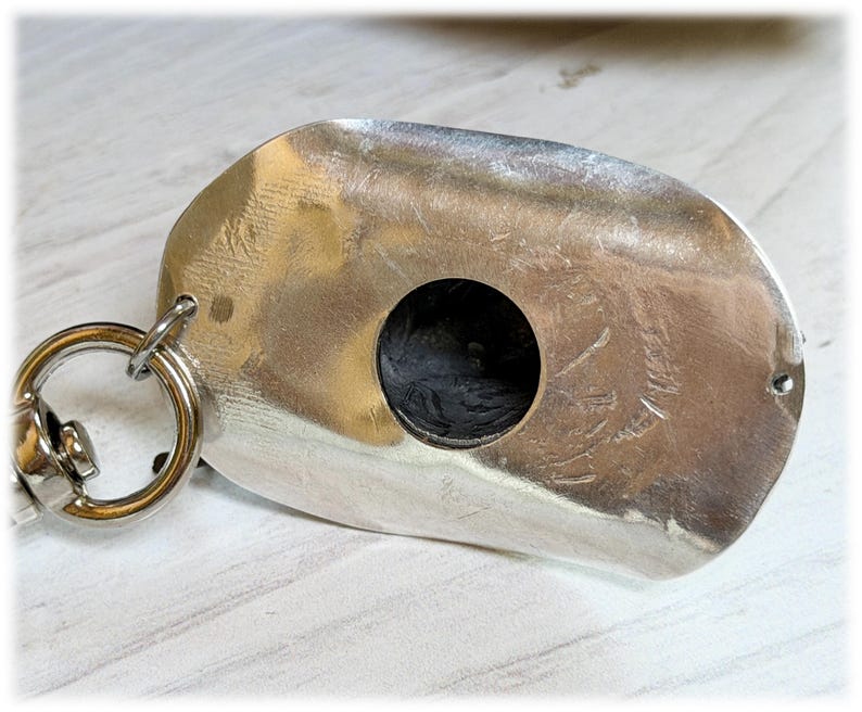 May include: A silver-toned metal dog tag with a smooth, reflective surface and a central circular cutout. The tag features a small hole on the right side and is attached to a circular metal split ring on the left side, creating a keychain component.