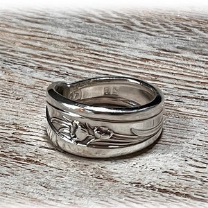 May include: A silver ring crafted from a spoon, featuring floral details. The ring has a polished finish and a band width of approximately 0.5 inches. The ring is displayed on a light-colored wooden surface, showcasing the intricate design and craftsmanship.