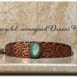 May include: Upcycled, reimagined drawer pull with a hammered copper finish and a light green glass cabochon.
