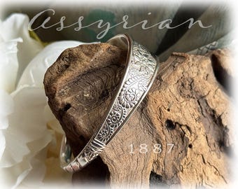 May include: A silver-colored bracelet with intricate floral and geometric patterns. The bracelet is curved and rests on a piece of wood. The word "Assyrian" is written in cursive script above the bracelet, and the number "1887" is below it.