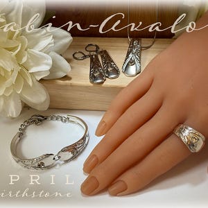 Puede incluir: Un brazalete y un anillo de plata hechos con cubiertos reutilizados con un diseño ornamentado. Las joyas se muestran en una mano con esmalte de uñas marrón claro. El texto "Cabin-Avalon" y "April Birthstone" es visible en la imagen.