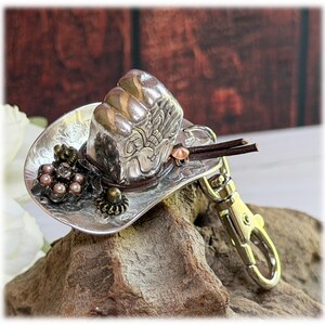 May include: A silver-toned metal cowboy hat keychain featuring an embossed floral pattern on the brim. The hat is decorated with a cluster of small metallic beads, a dark brown leather cord band, and a silver-colored metal lobster claw clasp.