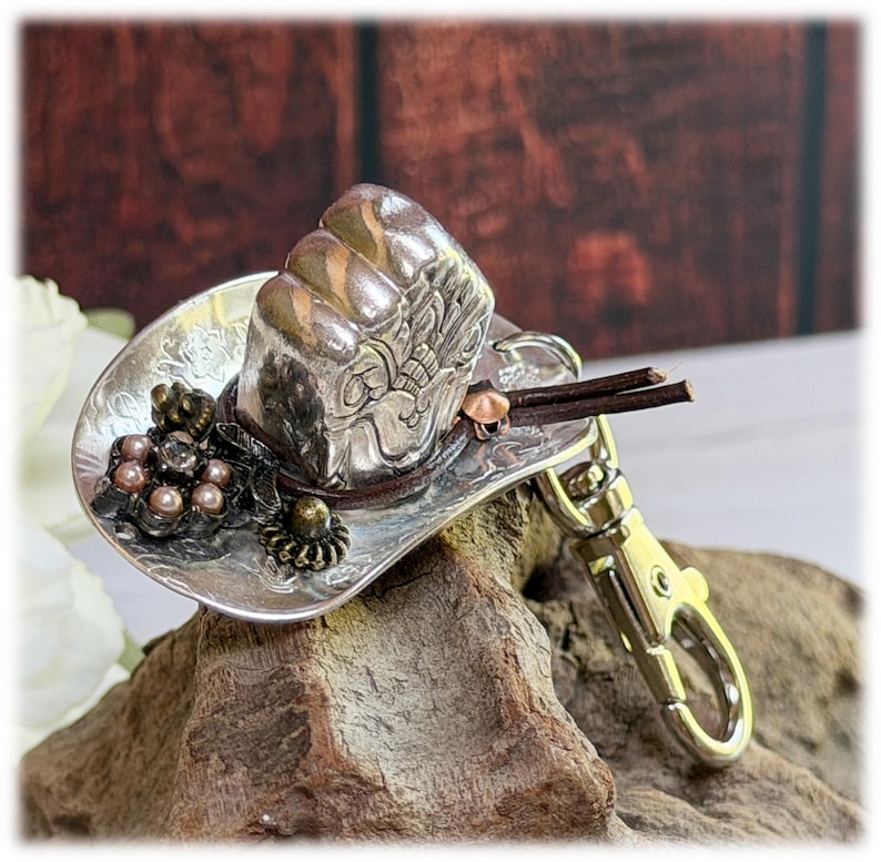 May include: A silver-toned metal cowboy hat keychain featuring an embossed floral pattern on the brim. The hat is decorated with a cluster of small metallic beads, a dark brown leather cord band, and a silver-colored metal lobster claw clasp.