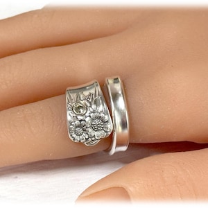 May include: A silver ring with a floral design and a small clear stone in the center. The ring is wrapped around a finger.