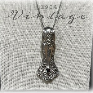 May include: A silver vintage spoon necklace with a dark purple gemstone pendant. The spoon has a grapevine design and the text "1904 Vintage" is visible in the background.