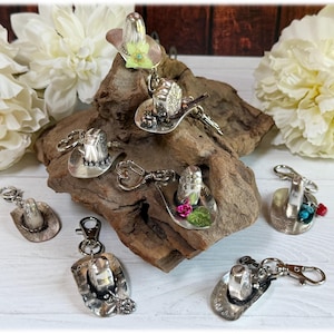 May include: A collection of silver-toned metal cowboy hat keychains, each featuring a unique decorative accent such as small flowers, turquoise-colored beads, or red stones, attached to a metal lobster clasp for secure attachment to items.