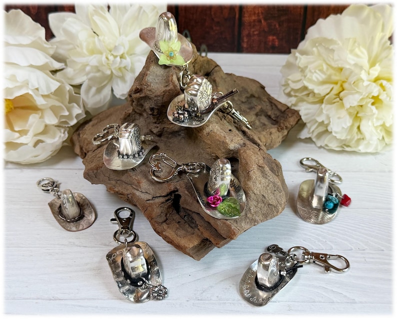 May include: A collection of silver-toned metal cowboy hat keychains, each featuring a unique decorative accent such as small flowers, turquoise-colored beads, or red stones, attached to a metal lobster clasp for secure attachment to items.