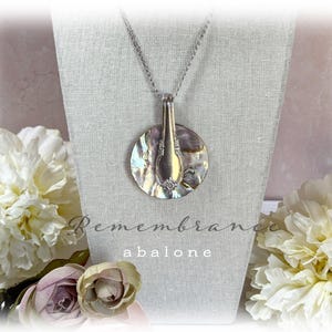 May include: A silver necklace with a pendant made from a vintage spoon and abalone shell. The pendant is round and has a iridescent finish. The necklace is on a white fabric background with the words "Remembrance abalone" printed below.