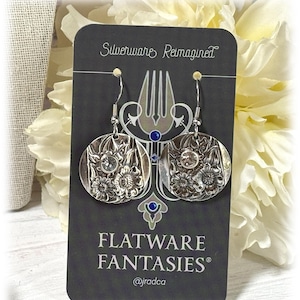 May include: Silver earrings with a floral design, made from repurposed silverware. The earrings are on a black card with the text "Silverware Reimagined" and "Flatware Fantasies" with the social media handle "@jradca".