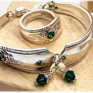 May include: A silver bracelet and ring set, made from repurposed silverware. The bracelet has decorative edges and is embellished with green gemstones. The ring has a matching design with a single green gemstone. The set is displayed on a wooden surface.