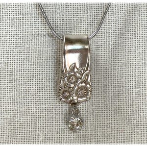 May include: A silver pendant necklace featuring a repurposed spoon handle. The pendant is adorned with floral detailing and a dangling clear crystal. The necklace hangs on a thin silver chain, set against a neutral textured background.