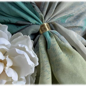 May include: A gold-colored scarf ring secures a folded scarf in shades of green and gray, with a floral pattern. White flowers are arranged on the left and right sides of the scarf, creating a soft, elegant composition. The scarf ring has an ornate design.