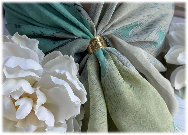 May include: A gold-colored scarf ring secures a folded scarf in shades of green and gray, with a floral pattern. White flowers are arranged on the left and right sides of the scarf, creating a soft, elegant composition. The scarf ring has an ornate design.