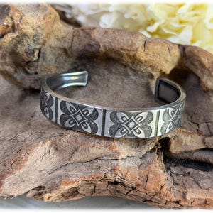 Puede incluir: Una pulsera de brazalete de metal plateado con un patrón repetitivo de motivos florales de cuatro pétalos separados por líneas verticales. La pulsera tiene un acabado pulido con áreas empotradas oscurecidas que resaltan el diseño grabado.