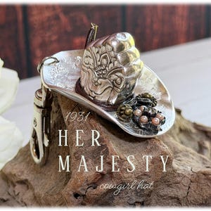May include: A silver-toned metal cowboy hat keychain featuring intricate embossed floral patterns on the brim and crown. The hat is adorned with a small cluster of dark metallic beads and light pink pearls. It includes a silver metal lobster clasp.