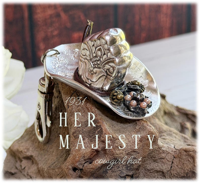 May include: A silver-toned metal cowboy hat keychain featuring intricate embossed floral patterns on the brim and crown. The hat is adorned with a small cluster of dark metallic beads and light pink pearls. It includes a silver metal lobster clasp.