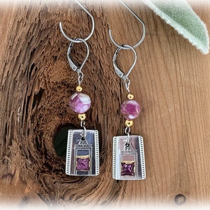May include: Silver and gold earrings with a purple and white bead and a square purple stone in a silver frame.