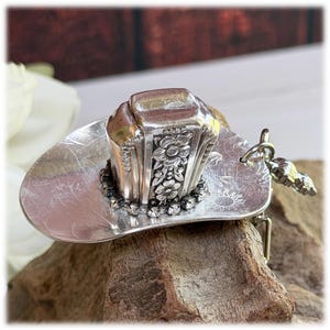 May include: A silver-toned metal cowboy hat ornament crafted from a repurposed spoon. The crown features an embossed floral pattern, while the brim is accented with a ring of small clear rhinestones and a small engraved sun symbol on the side.