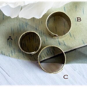 May include: Three gold-colored adjustable rings are displayed on a patterned fabric. The rings are circular with a textured surface. The rings are labeled A, B, and C.