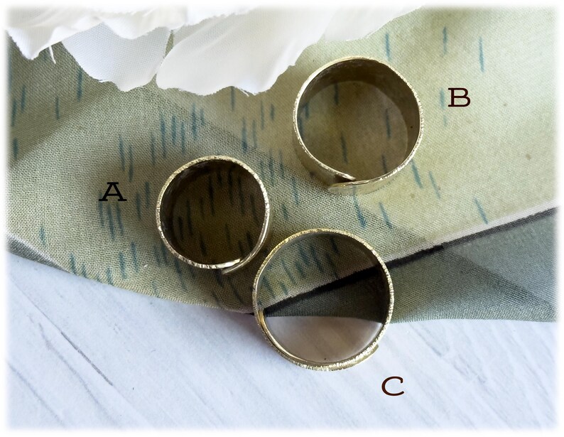 May include: Three gold-colored adjustable rings are displayed on a patterned fabric. The rings are circular with a textured surface. The rings are labeled A, B, and C.