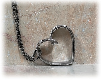 May include: A silver-toned heart pendant necklace. The open heart design is suspended from a dark chain. The necklace is displayed against a neutral, textured background.