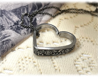 Könnte beinhalten: Halskette mit herzförmigem Anhänger in Silber. Der Anhänger hat ein florales Muster an den Rändern und hängt an einer feinen Kette. Die Kette liegt auf einer cremefarbenen Spitze, im Hintergrund ist ein gemusterter Stoff zu sehen.