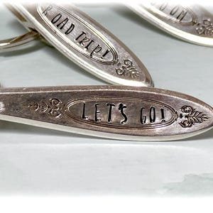 May include: Silver keyrings with stamped text. One reads "ROAD TRIP!" and another "LET'S GO!". The keyrings have a spoon-like shape with decorative floral accents. The background is a light blue colour.