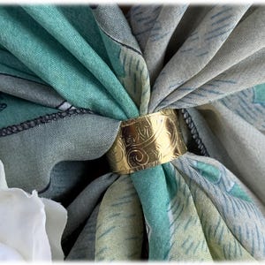 May include: Close-up of a teal and gray scarf held together by a gold-colored ring. The ring features an ornate, swirling design. The scarf has a subtle pattern and is draped to create a layered effect, with a white flower in the corner.
