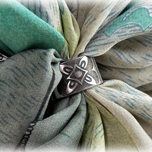 Könnte beinhalten: Ein silberner Schalring mit floralem Design ist auf einem gefalteten Schal zentriert. Der Schal ist eine Mischung aus grünen, blauen und cremefarbenen Farben mit einem dezenten Muster. Der Ring hat eine strukturierte Oberfläche und ein detailliertes Design.