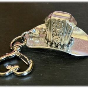 May include: A silver-toned metal keychain featuring a miniature cowboy hat charm. The hat is decorated with embossed floral patterns on the crown, a row of small clear rhinestones around the base, and a heart-shaped lobster claw clasp attachment.