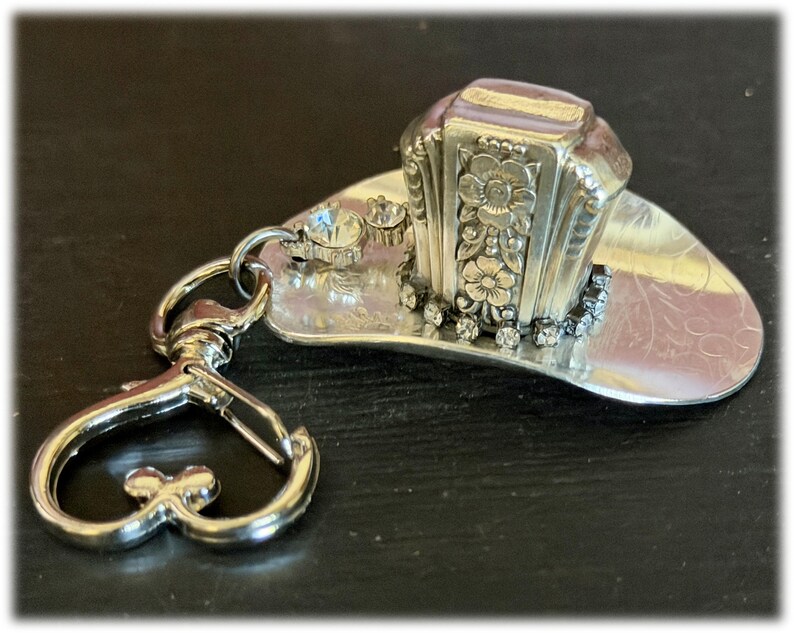 May include: A silver-toned metal keychain featuring a miniature cowboy hat charm. The hat is decorated with embossed floral patterns on the crown, a row of small clear rhinestones around the base, and a heart-shaped lobster claw clasp attachment.