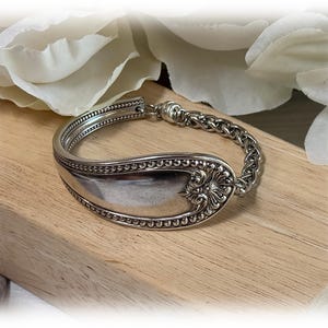 Könnte beinhalten: Ein silbernes Armband aus einem Löffel, mit aufwendigen floralen und perlenbesetzten Details. Die Löffelschale bildet den Hauptteil, verbunden mit einem Kettengliedband. Das Armband liegt auf einer Holzoberfläche, mit weißen Blumen im Hintergrund.