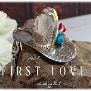 May include: A silver-toned metal keychain shaped like a cowboy hat with embossed floral patterns. The hat is adorned with small turquoise and red beads. The text 1937 FIRST LOVE cowboy hat is visible on the image, along with a metal lobster clasp.