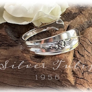 May include: A silver spoon bracelet with floral details, resting on a piece of wood. The bracelet is engraved with the words "Silver Tulip" and the year "1956". A white flower is in the background.