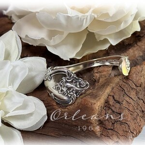 May include: A decorative key made from a silver spoon, with intricate floral patterns on the spoon's bowl. The key rests on a piece of aged wood, surrounded by white fabric flowers. The word "Orleans" and the number "1964" are visible.
