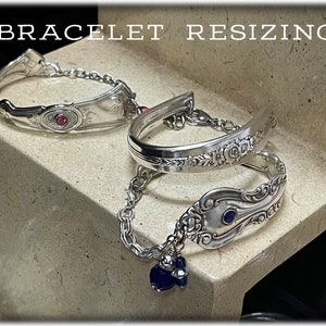 May include: Two silver bracelets with decorative designs and gemstones. The bracelet on the left has a pink gemstone, while the bracelet on the right has a blue gemstone. The bracelets are on a light brown surface.