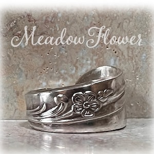 May include: A silver cuff bracelet with a floral design. The bracelet has a curved shape and features a raised flower and leaf pattern. The text "Meadow Flower" is above the bracelet.