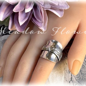 Può includere: Anello in argento fatto a mano con un design unico. L'anello è indossato su un dito, con la fascia che avvolge il dito. Un fiore viola è vicino. Il testo "Meadow Flower" è sullo sfondo.