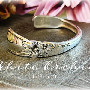 May include: A silver-toned metal cuff bracelet crafted from a repurposed spoon handle, featuring an embossed floral pattern with petals and leaves. The text White Orchid 1953 is displayed in white lettering below the curved metal accessory.