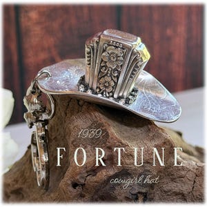 May include: A silver-toned metal ornament shaped like a cowboy hat, crafted from a repurposed spoon. The hat features intricate floral engravings on the crown and a curved brim. The text 1939 FORTUNE cowgirl hat is visible on the image.