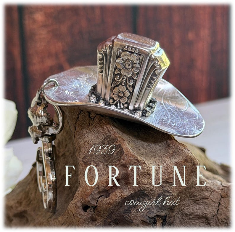 May include: A silver-toned metal ornament shaped like a cowboy hat, crafted from a repurposed spoon. The hat features intricate floral engravings on the crown and a curved brim. The text 1939 FORTUNE cowgirl hat is visible on the image.