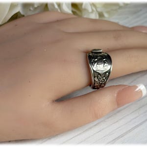 May include: A silver-toned metal ring crafted from a repurposed spoon handle. The band features an embossed crest at the top and a detailed floral pattern along the lower section, with a polished finish that highlights the intricate metalwork.