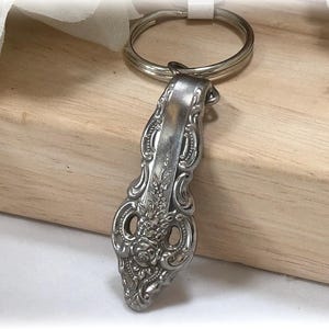 Puede incluir: Llavero plateado en forma de cuchara ornamentada. El mango de la cuchara presenta un diseño floral con detalles intrincados. El llavero incluye un anillo de metal plateado para sujetar llaves. La cuchara es de metal brillante y reflectante.