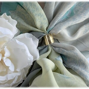 May include: A close-up of a silk scarf with a gold-colored ring clasp. The scarf features a pattern of light blue, green, and cream colors. White flowers are in the foreground.
