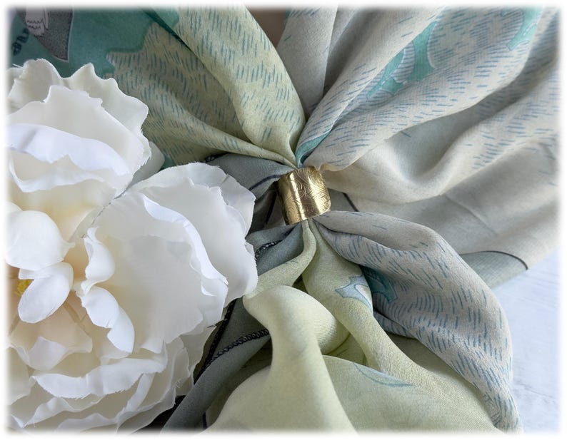 May include: A close-up of a silk scarf with a gold-colored ring clasp. The scarf features a pattern of light blue, green, and cream colors. White flowers are in the foreground.