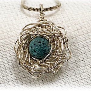 May include: A silver pendant necklace with a nest-shaped design. The nest is made from woven wire, holding a teal-coloured, spherical stone. The chain is a delicate, textured silver. The pendant hangs against a white, textured background.