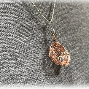 Könnte beinhalten: Eine kupferfarbene, drahtgewickelte Anhänger-Halskette. Der Anhänger ist ein gewebtes, tropfenförmiges Design, das an einer filigranen Silberkette hängt. Die Halskette wird vor einem grauen Hintergrund präsentiert.
