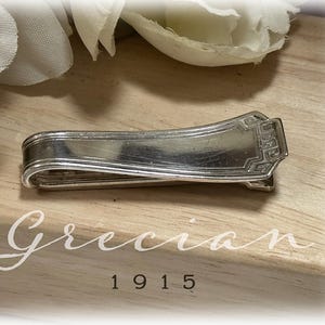 May include: A silver-colored tie clip with a decorative rectangular end. The clip is curved and has a simple design. The word "Grecian" and the number "1915" are printed on the wooden surface beneath the clip.