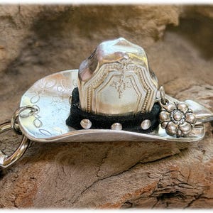 May include: A silver-toned metal cowboy hat keychain featuring a curved brim with engraved patterns. The crown is adorned with a black fabric band accented by small silver studs and a dangling silver flower charm attached to the side.