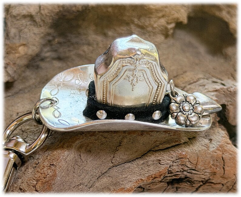 May include: A silver-toned metal cowboy hat keychain featuring a curved brim with engraved patterns. The crown is adorned with a black fabric band accented by small silver studs and a dangling silver flower charm attached to the side.